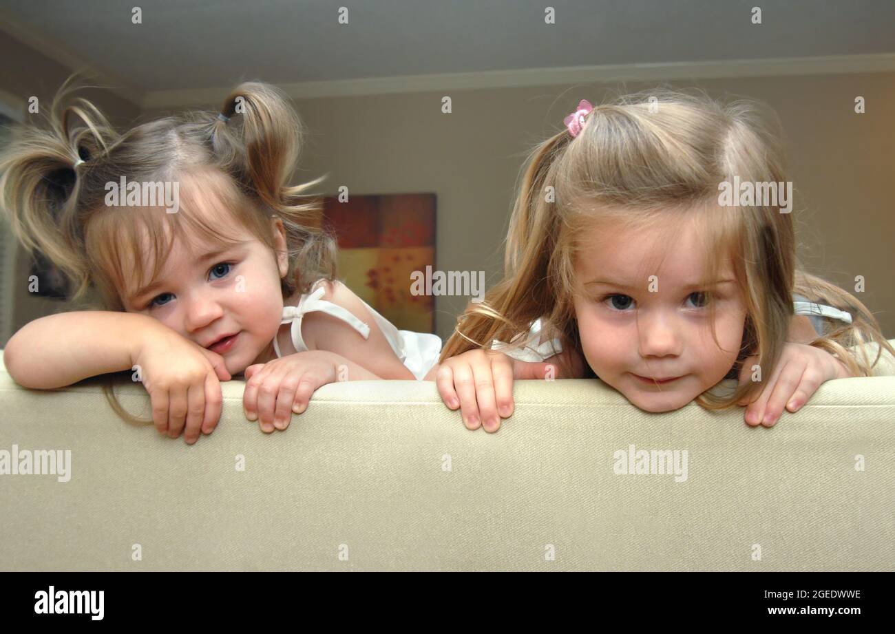 Zwei entzückende kleine Mädchen schauen über die Oberseite des Sofas in ihrem Haus. Schwestern, diese beiden sehen aus wie Ärger. Der jüngste hat Zöpfe. Stockfoto
