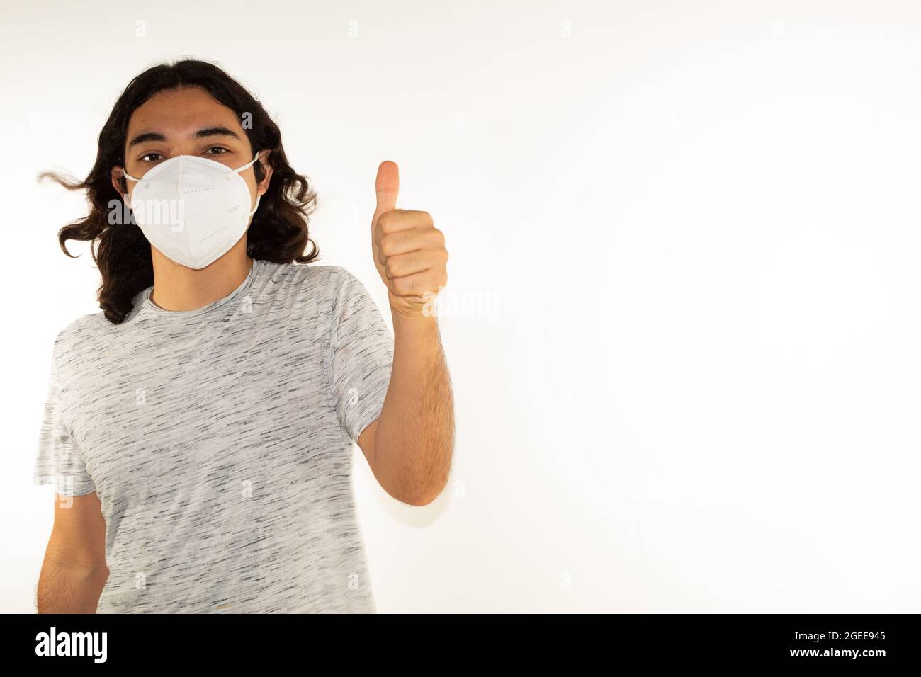 Hispanic Mann mit langen Haaren trägt eine kn95 Gesichtsmaske auf weißem Hintergrund mit einem Daumen nach oben Stockfoto