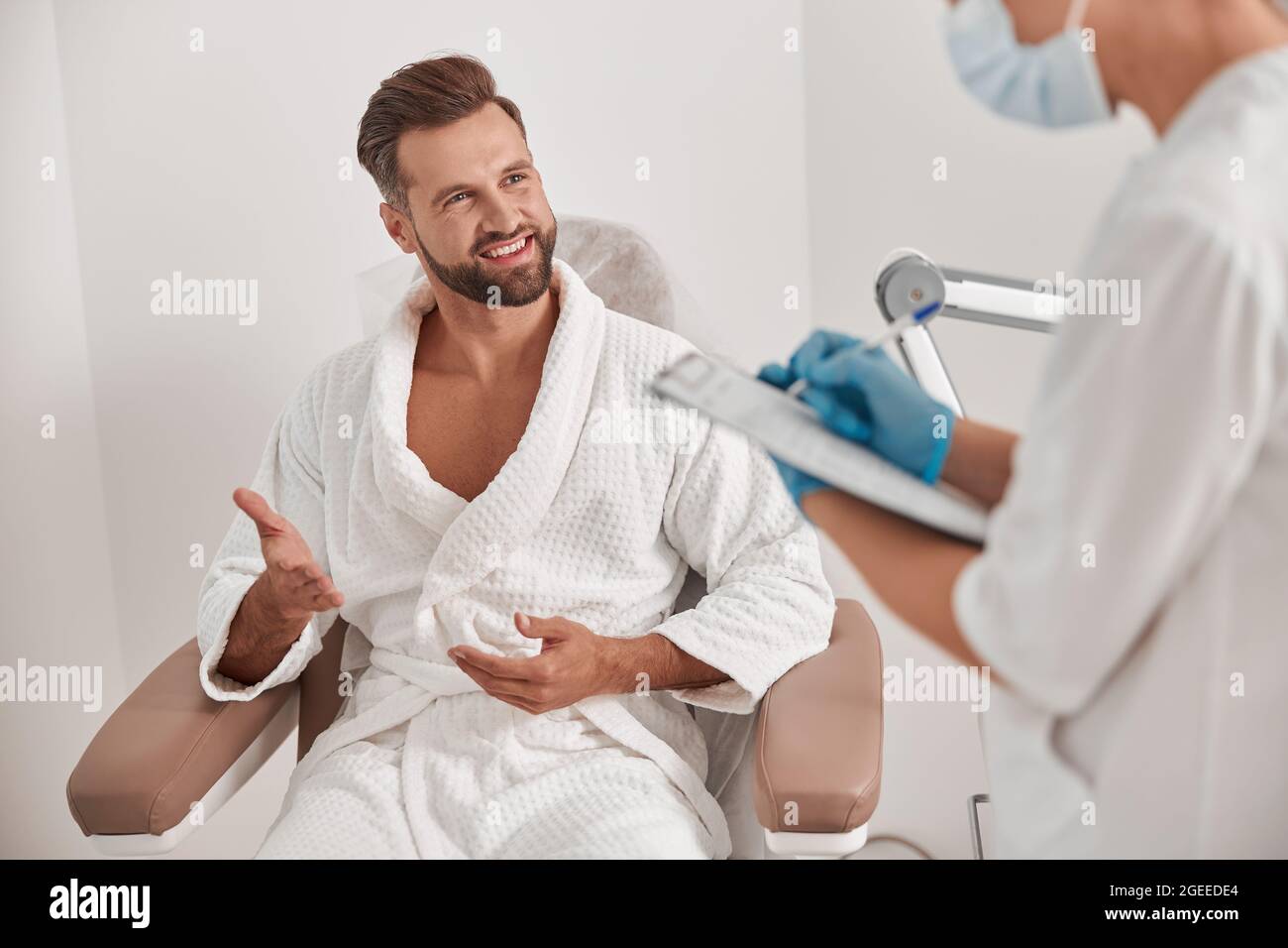 Lächelnder Mann spricht mit dem Administrator, der das Formular ausfüllt, während er den Patienten in der Klinik untersucht Stockfoto