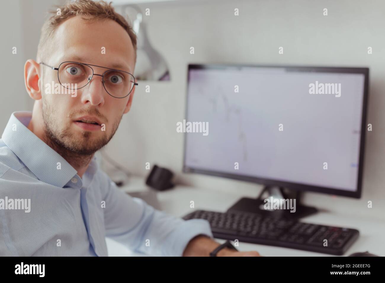 Porträt eines überraschten Mannes in Brille und Bürohemd auf dem Hintergrund des Monitors mit Grafik des fallenden Marktes. Börsenkrise. Kryptowährung. Lott Stockfoto