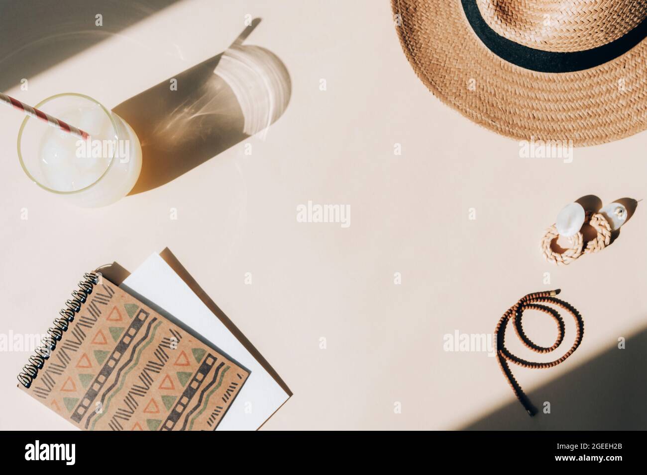 Schreibtisch mit Notizblock, frischer Limonade mit Eiswürfeln, Hut und Ohrringen auf beigefarbenem Hintergrund. Sommerferienkonzept, Draufsicht, Flat Lay, Copy Space. Stockfoto
