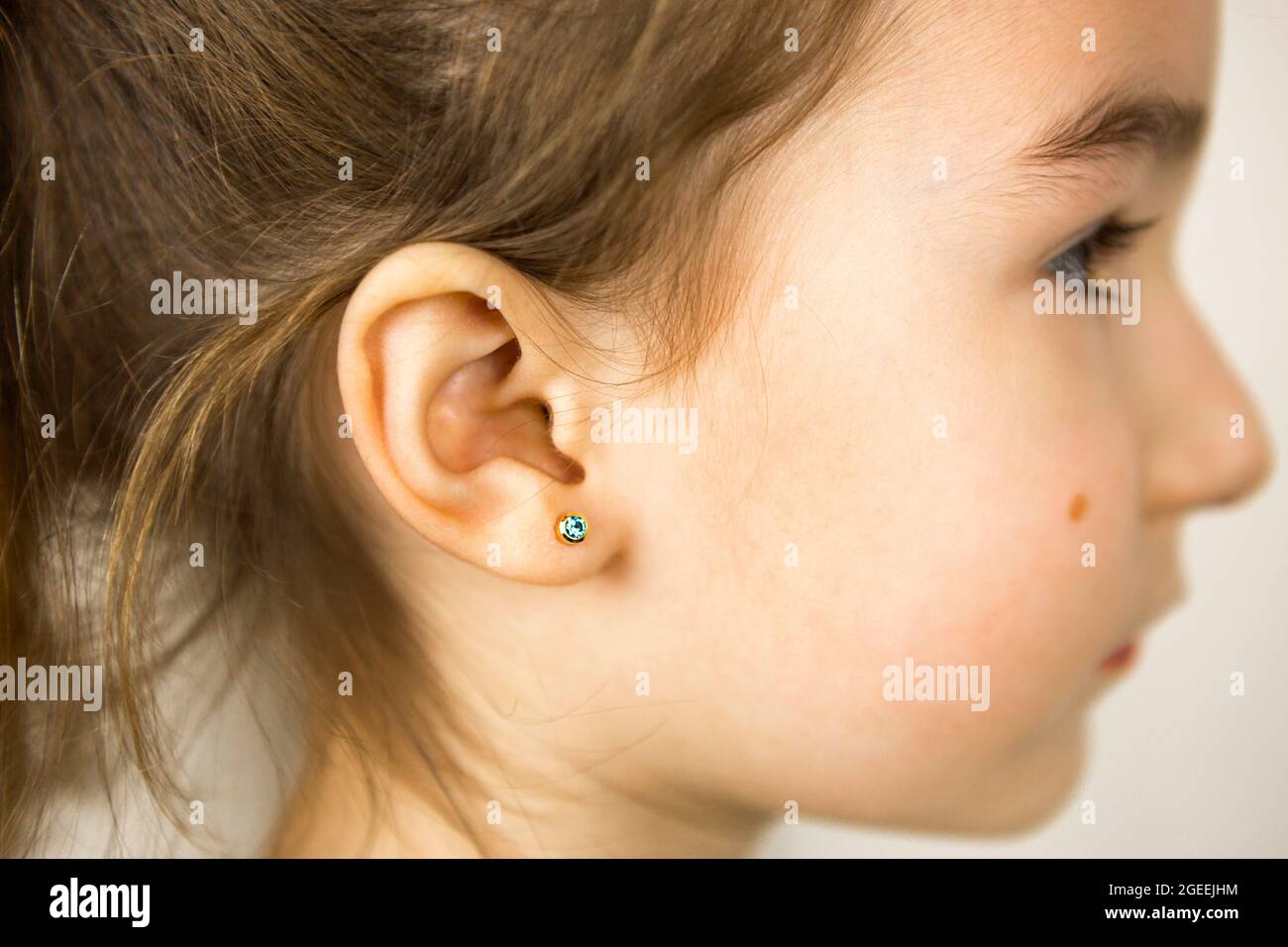 Ohrpiercing bei einem Kind - ein Mädchen zeigt einen Ohrring in ihrem Ohr aus einer medizinischen Legierung. Weißer Hintergrund, Porträt eines Mädchens mit einem Maulwurf auf der Wange Stockfoto