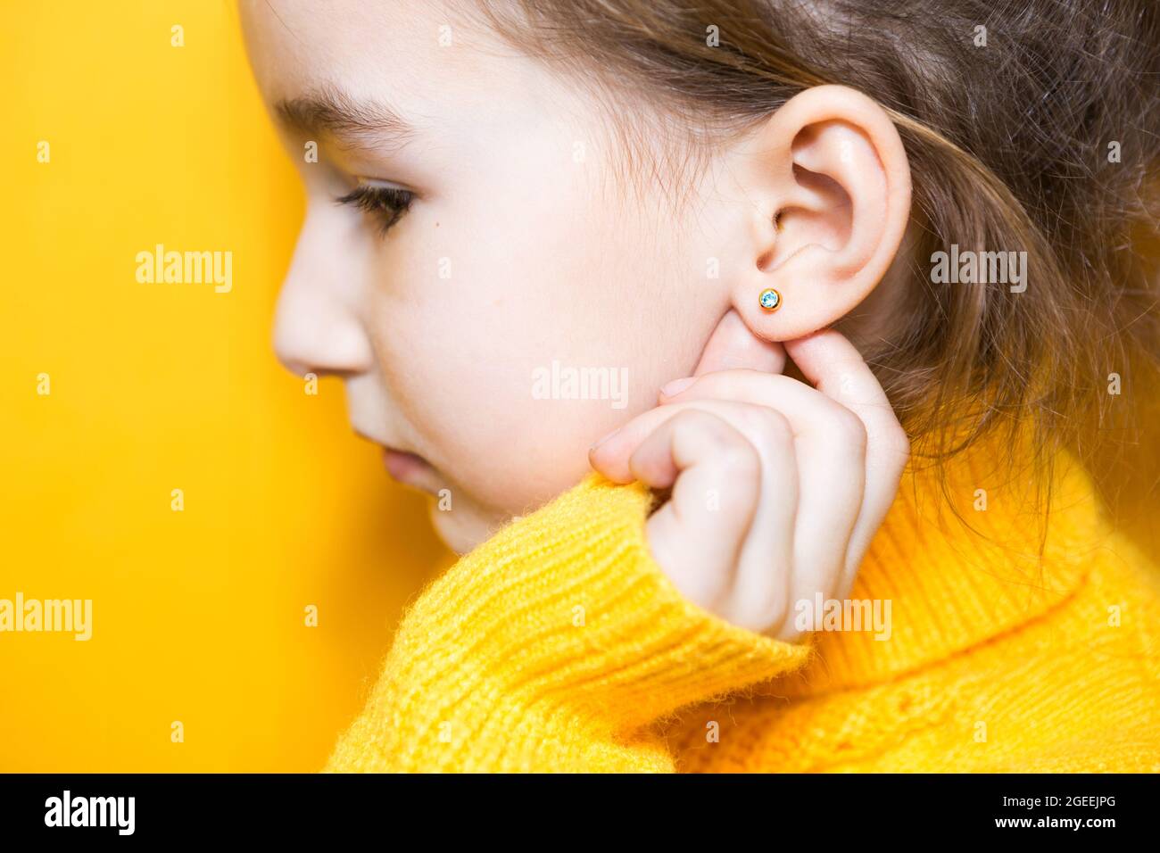 Ohrpiercing bei einem Kind - ein Mädchen zeigt einen Ohrring in ihrem Ohr aus einer medizinischen Legierung. Gelber Hintergrund, Porträt eines Mädchens im Profil. Stockfoto