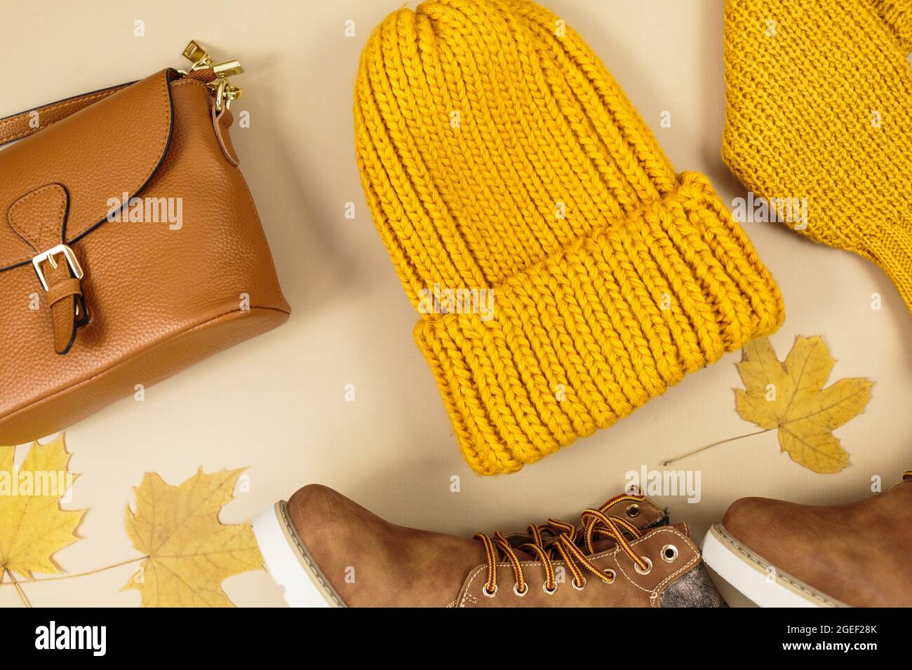 Orangefarbene Ledertasche, Strickpullover, Hut, Herbstblätter auf beigem Hintergrund. Trendige Herbst-Accessoires. Gemütliches Outfit Stockfoto