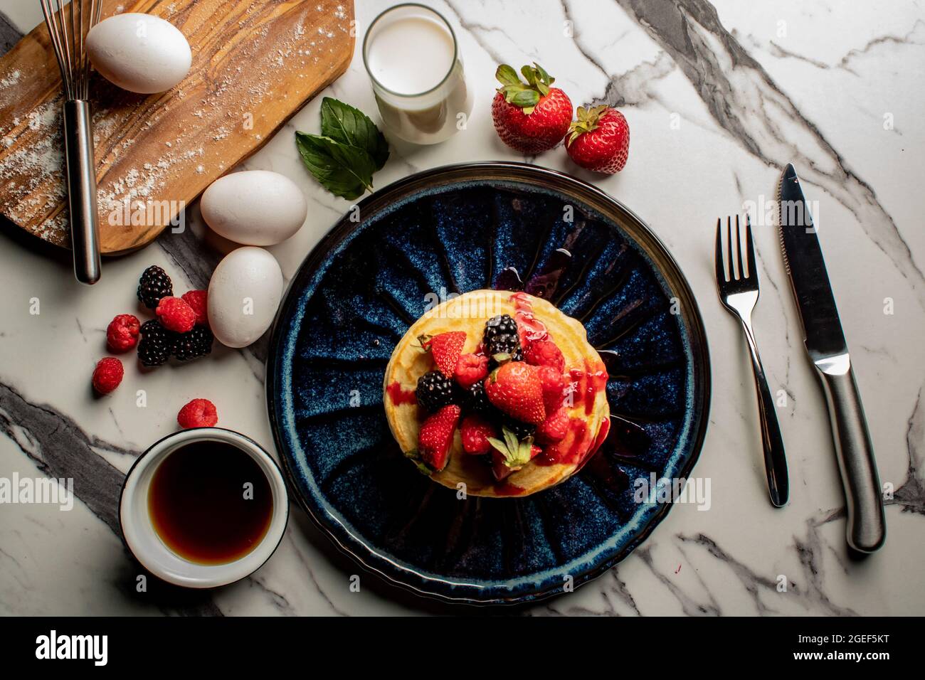 Ein Stapel appetitanregende Pfannkuchen mit frischen Beeren, Zutaten und Utensilien auf einer Marmoroberfläche Stockfoto