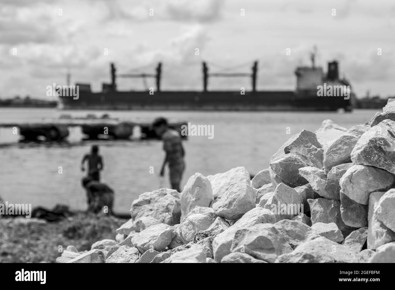 Graustufenaufnahme von gestapelten Felsen am Ufer mit Kindern, die in der Nähe des Meeres im verschwommenen Hintergrund spielen Stockfoto