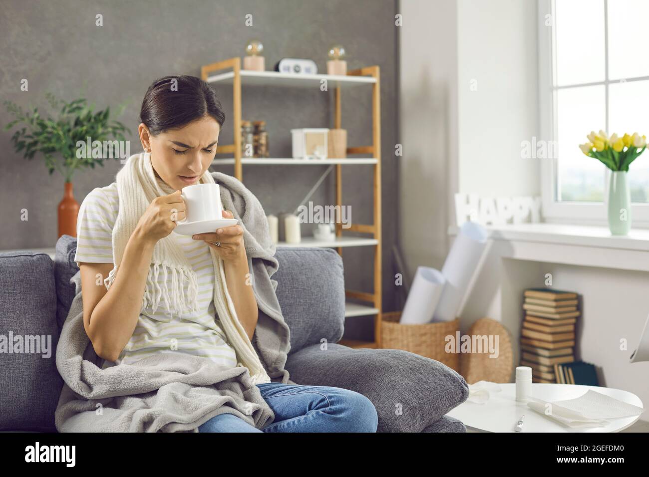 Eine Frau, die an einer Erkältung oder Grippe mit Fieber leidet, trinkt warmen Tee, während sie zu Hause auf der Couch sitzt Stockfoto