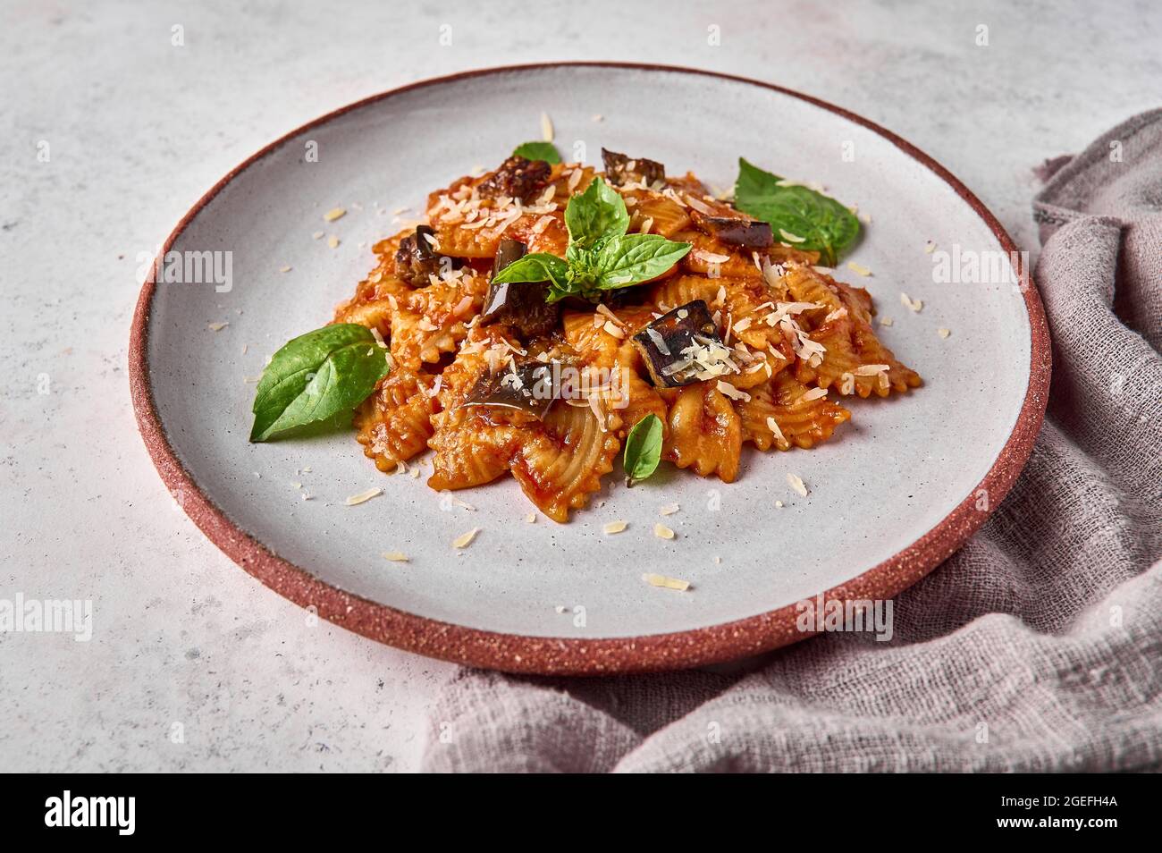 Farfalle Pasta mit Auberginen, Tomatensauce, Parmesan und Basilikum auf Teller mit Serviette auf grauem Hintergrund, Nahaufnahme. Selektiver Fokus Stockfoto
