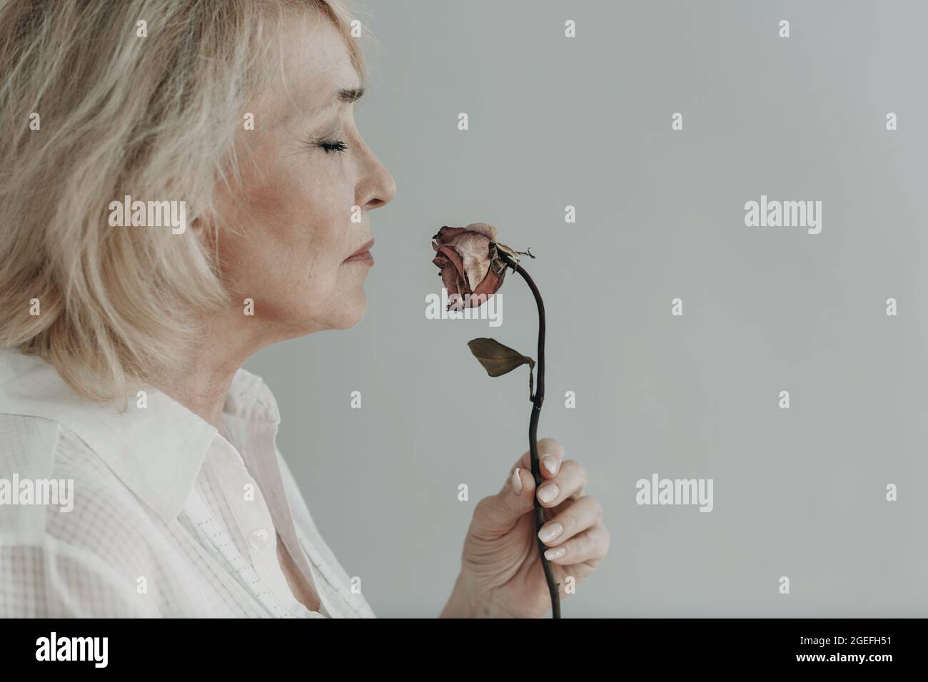 Traurige ältere Frau im weißen Hemd hält eine trockene graue alte tote Rosenblume. Altes alterndes Konzept. Stockfoto