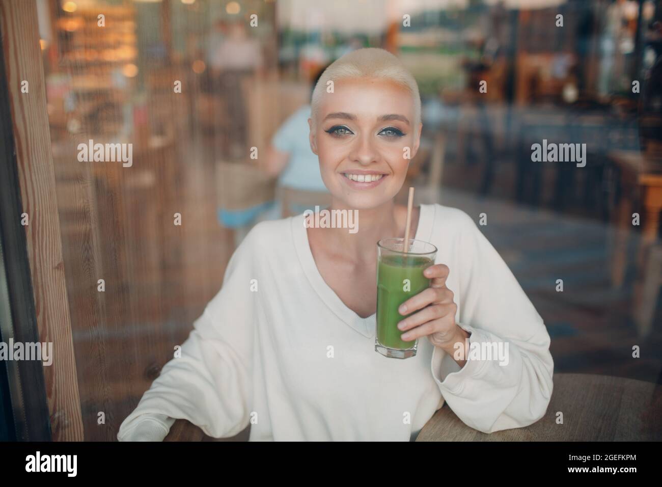 Porträt einer jungen, lächelnden, tausendjährigen europäischen Kurzhaarfrau, die mit grünem Smoothie im Café durch Glas schaut. Schöne glücklich blonde Mädchen innen. Stockfoto