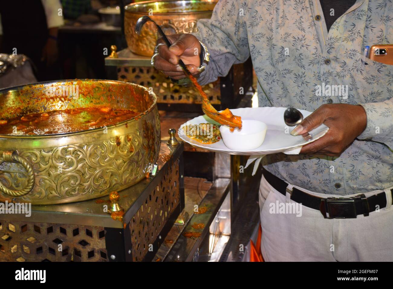 Indischer Mann, der Paneer Gemüse in seinem Teller aus einem hellen großen Topf bei der Hochzeitszeremonie trägt Stockfoto