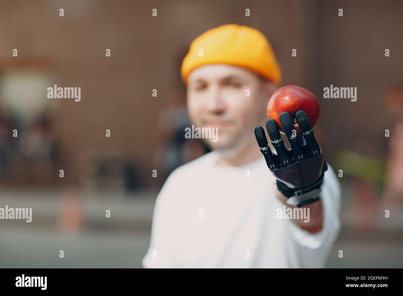 Ein junger behinderter Mann hält dir einen Apfel mit einer künstlichen prothetischen Hand in legerer Kleidung aus Stockfoto