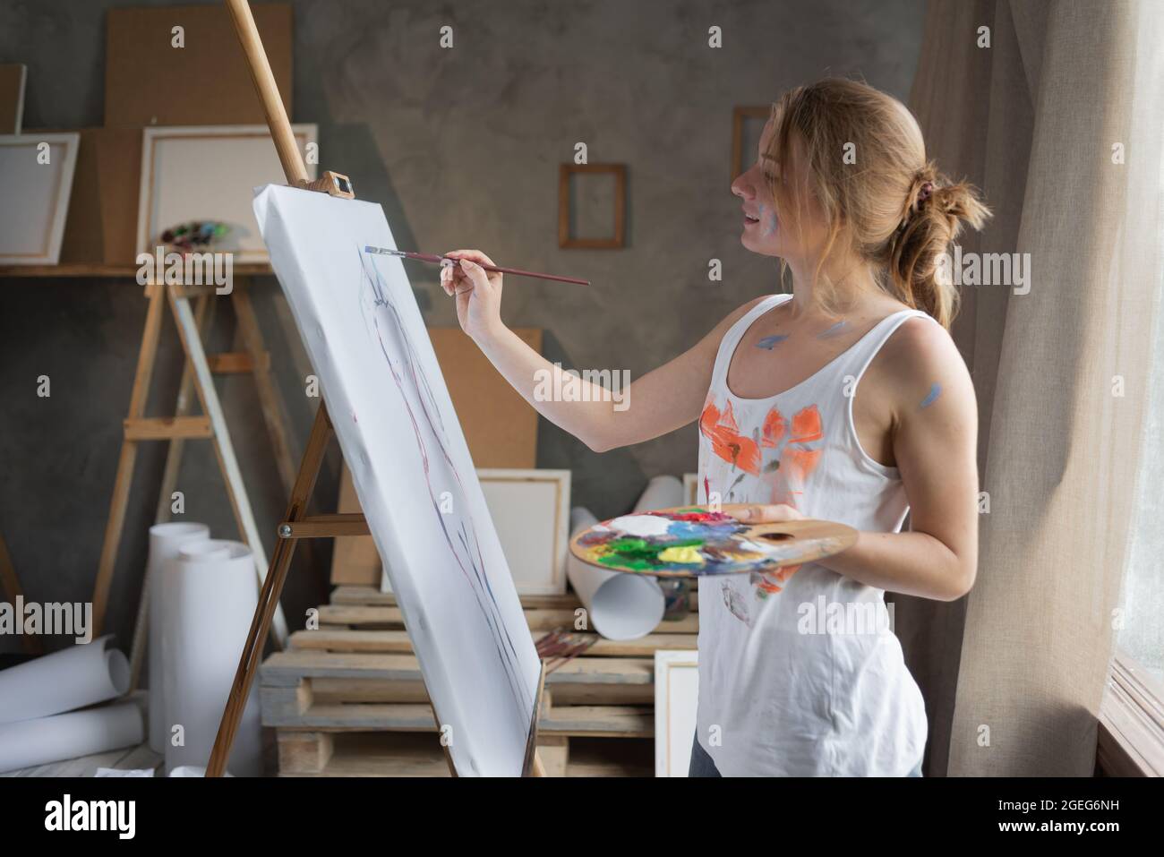 Eine kaukasische Künstlerin in einem schmutzigen T-Shirt steht in der Nähe einer Staffelei und trägt Farbstriche auf die Leinwand. Lieblings-kreatives Hobby-Konzept. Stockfoto