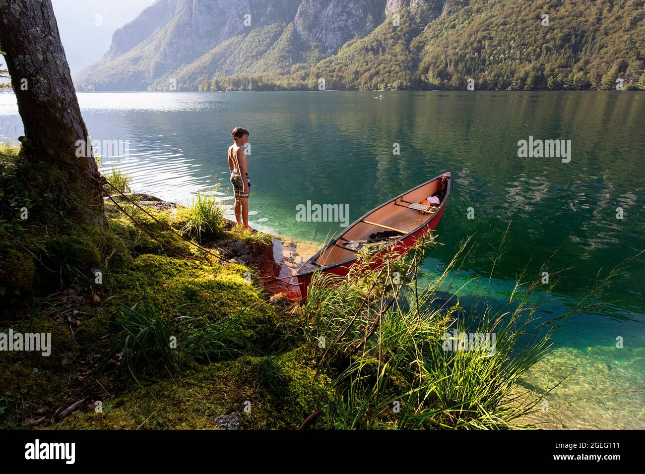 Ein Junge steht an einem heißen Sommertag während der Schulferien in slowenien am Ufer des wunderschönen Bohinj-Sees in der Nähe eines roten Kanus Stockfoto