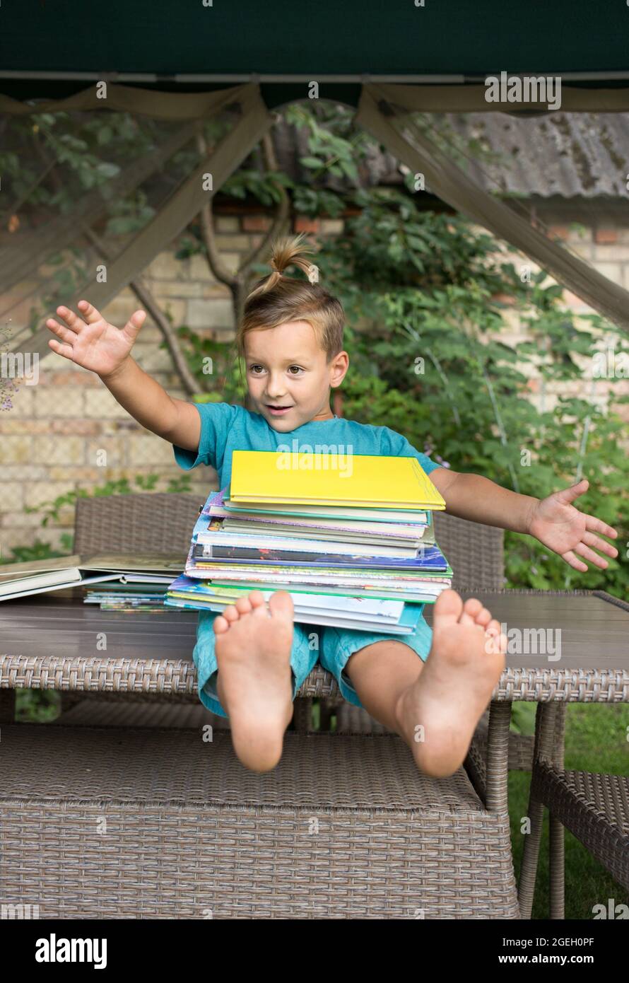Lustig fröhlich wissbegierig Vorschüler sitzt auf dem Tisch, hält einen großen Stapel von Büchern. Bücher lesen, Kinderfantasien, interessante Kindheit. Ins Stockfoto