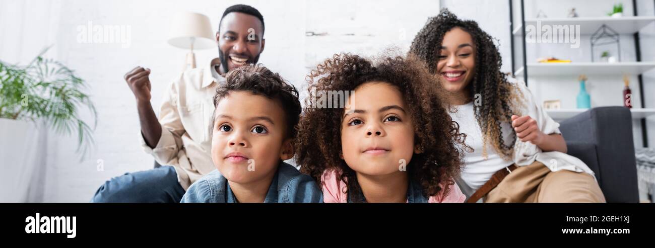 Konzentrierte afroamerikanische Geschwister, die in der Nähe von fröhlichen Eltern Fernsehen, zeigen eine Geste des Sieges und ein Banner Stockfoto