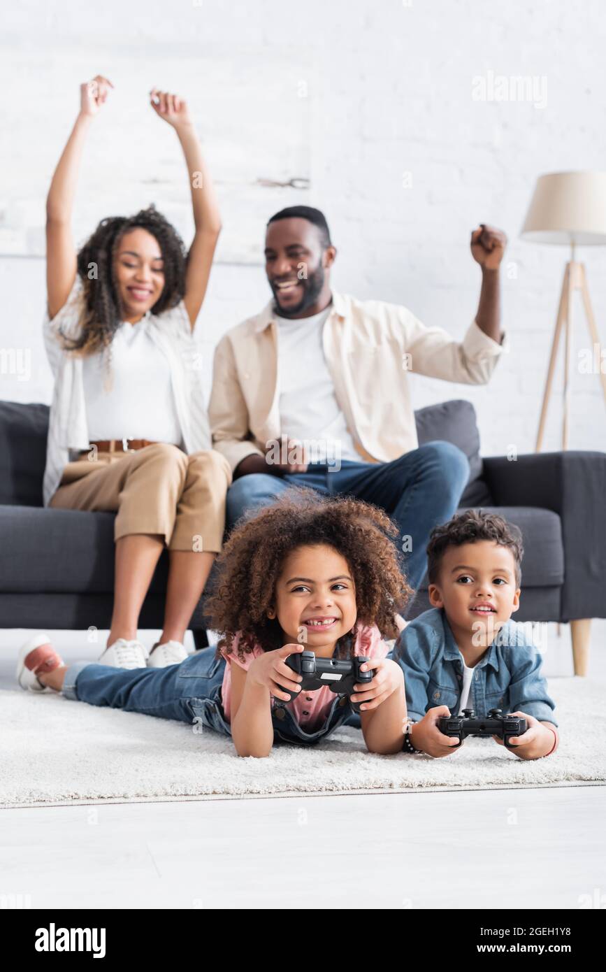 KIEW, UKRAINE - 25. JANUAR 2021: Aufgeregte afroamerikanische Eltern zeigen eine Siegestur in der Nähe von Kindern, die auf dem Boden spielen Stockfoto