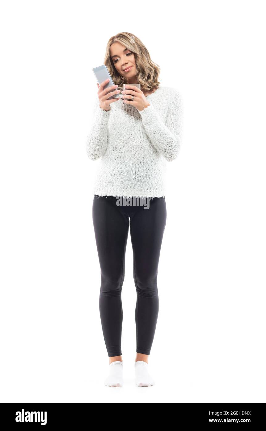 Nette junge Frau, die auf einem Smartphone im Internet surft und Kaffeebecher in gemütlicher Kleidung hält. Ganzkörperporträt isoliert auf weißem Hintergrund Stockfoto