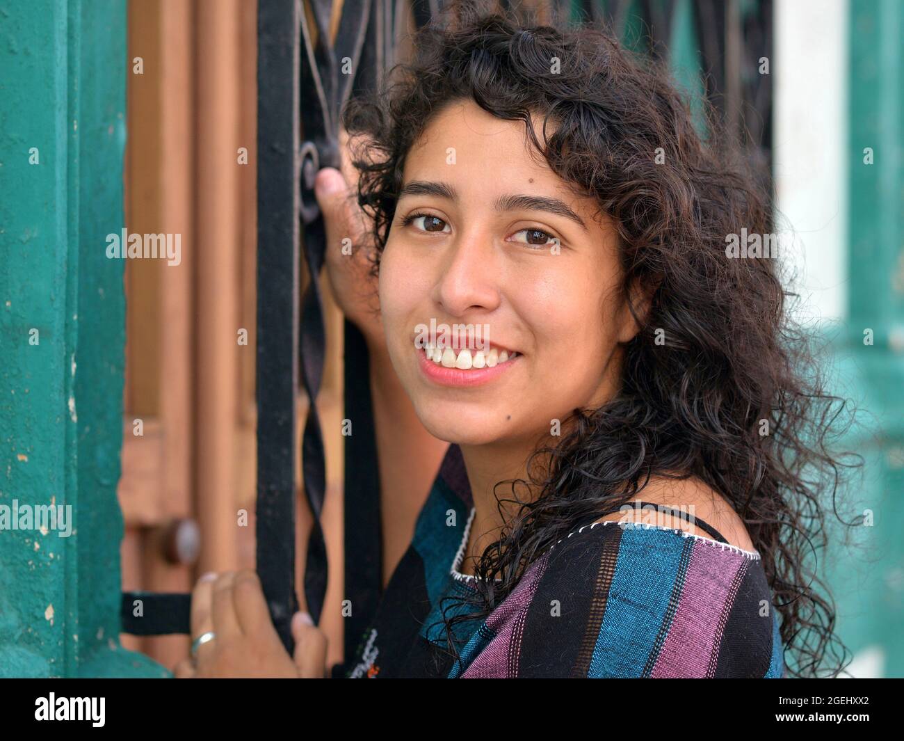 Schöne positive emotionale junge mexikanische Latina Hispanic Frau mit langen schwarzen Locken hält an einem schwarz lackierten Sicherheitsstahl Fenstergrill. Stockfoto
