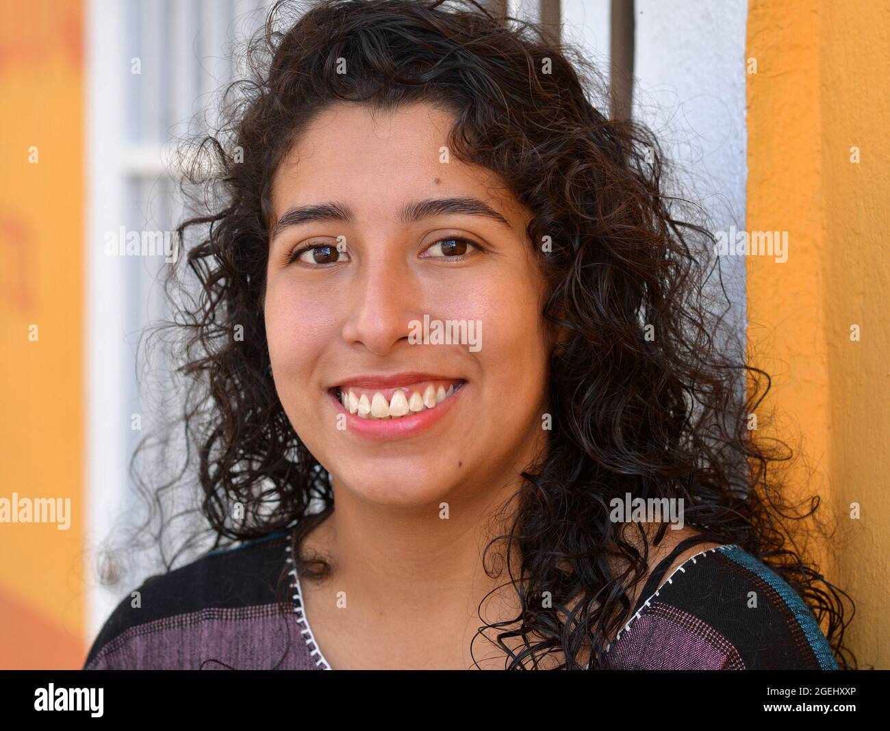 Schöne positive emotionale junge mexikanische Latina hispanische Frau mit langen schwarzen Locken lächelt vor einer weißen Sicherheitstür. Stockfoto