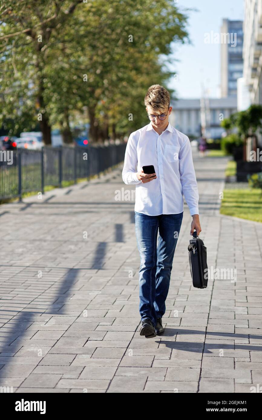 Junger Mann, der die Straße entlang läuft, mit einem Telefon in der Hand und einer Aktentasche in der anderen Hand. Geringer Fokus. Stockfoto