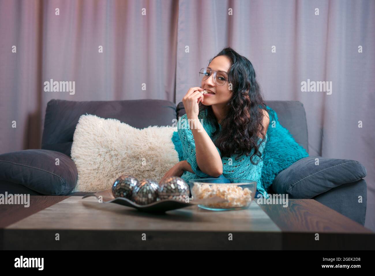 Horizontale Ansicht einer Frau, die zu Hause einen Film sieht, sitzt sie sehr ruhig auf ihrem Sofa und blickt auf die Kamera und isst viel Popcorn Stockfoto