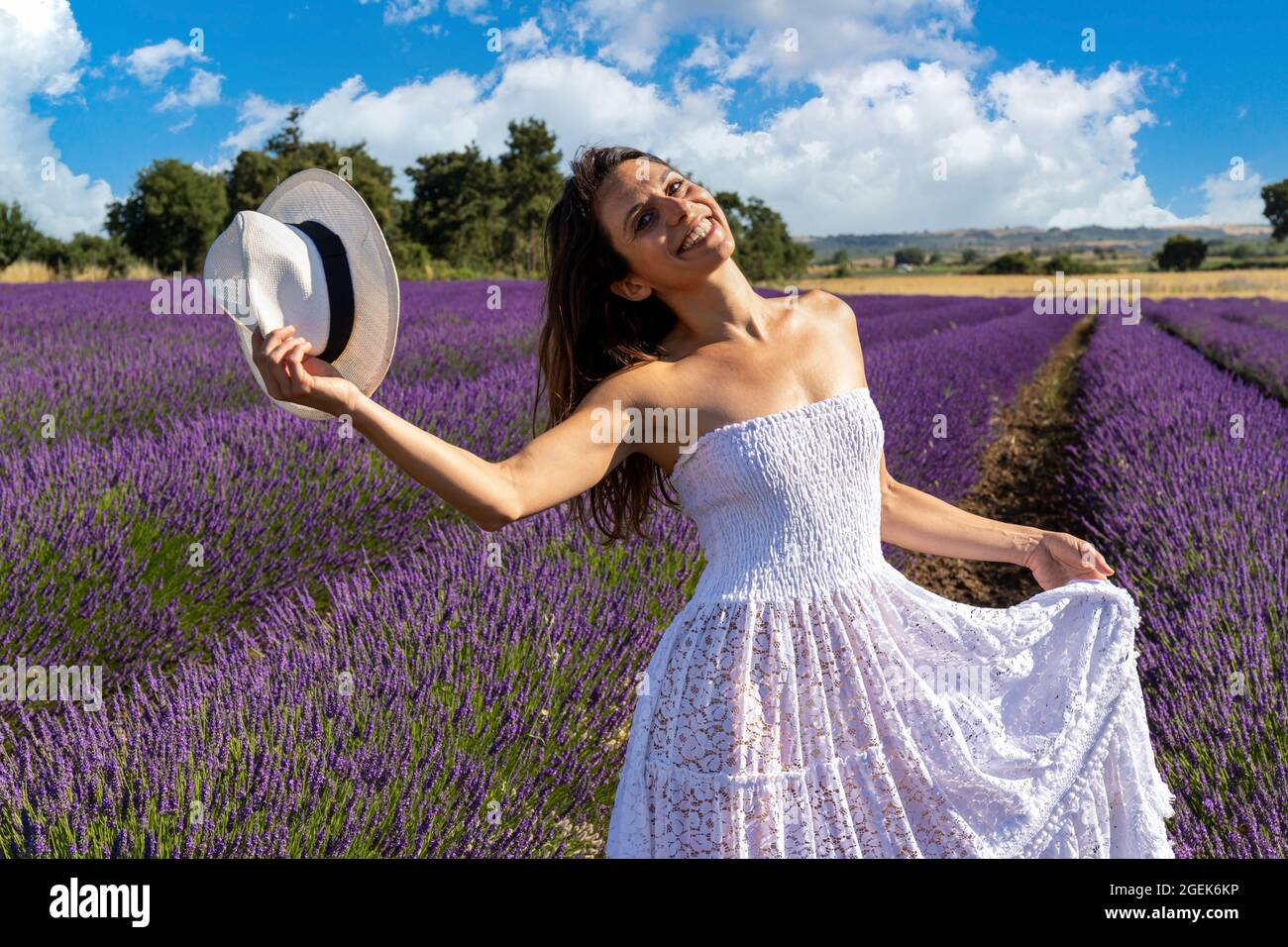 Porträt einer glücklichen jungen Frau, die mit ihrem Hut in einem blühenden Lavendelfeld spielt. Ihr weißes Kleid hebt sich von der violetten Farbe des Lavendelflos ab Stockfoto