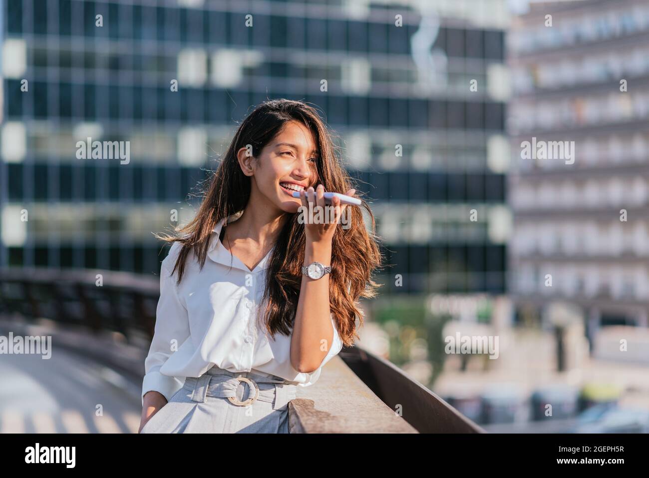 Horizontales Porträt einer attraktiven indischen Frau, die mit ihrem Smartphone eine Sprachnachricht sendet. Sie lehnt sich an das Geländer einer Brücke Stockfoto