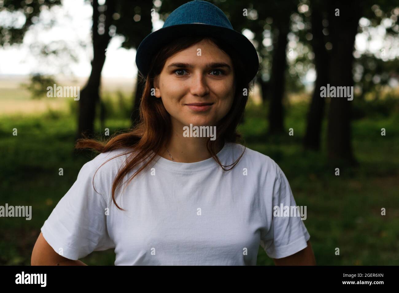 Unschärfe-Nahaufnahme-Porträt einer fröhlich lächelnden jungen Frau mit braunen Haaren, die einen Hut im Freien trägt. Grüne Natur Hintergrund. Blick auf die Kamera. Frauen Stockfoto