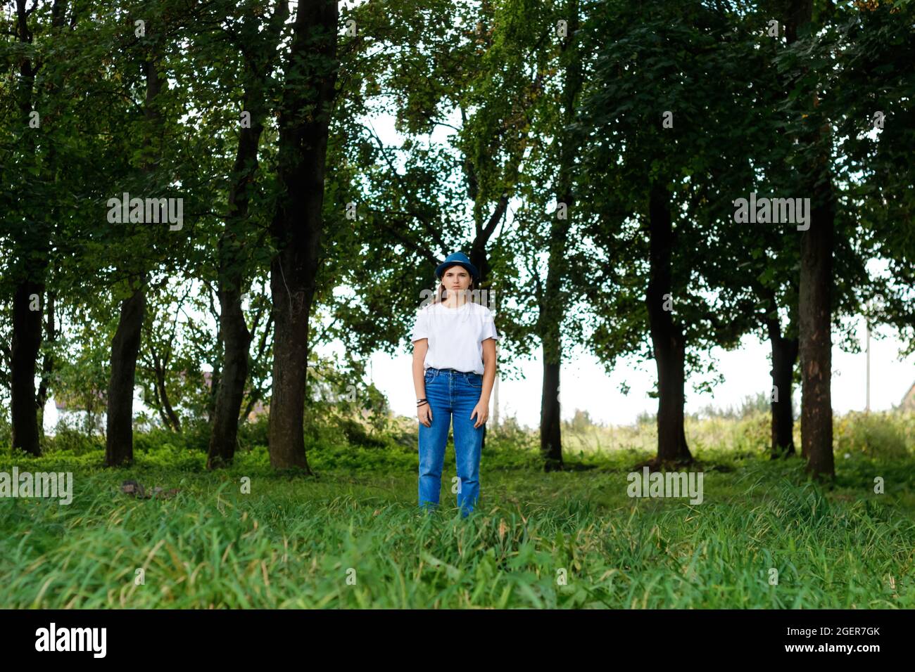 Unschärfe-Gesamtporträt einer ernsthaften jungen Frau mit braunen Haaren, die einen Hut im Freien trägt. Grüne Natur Bäume Hintergrund, Wald. Blick auf die Kamera Stockfoto