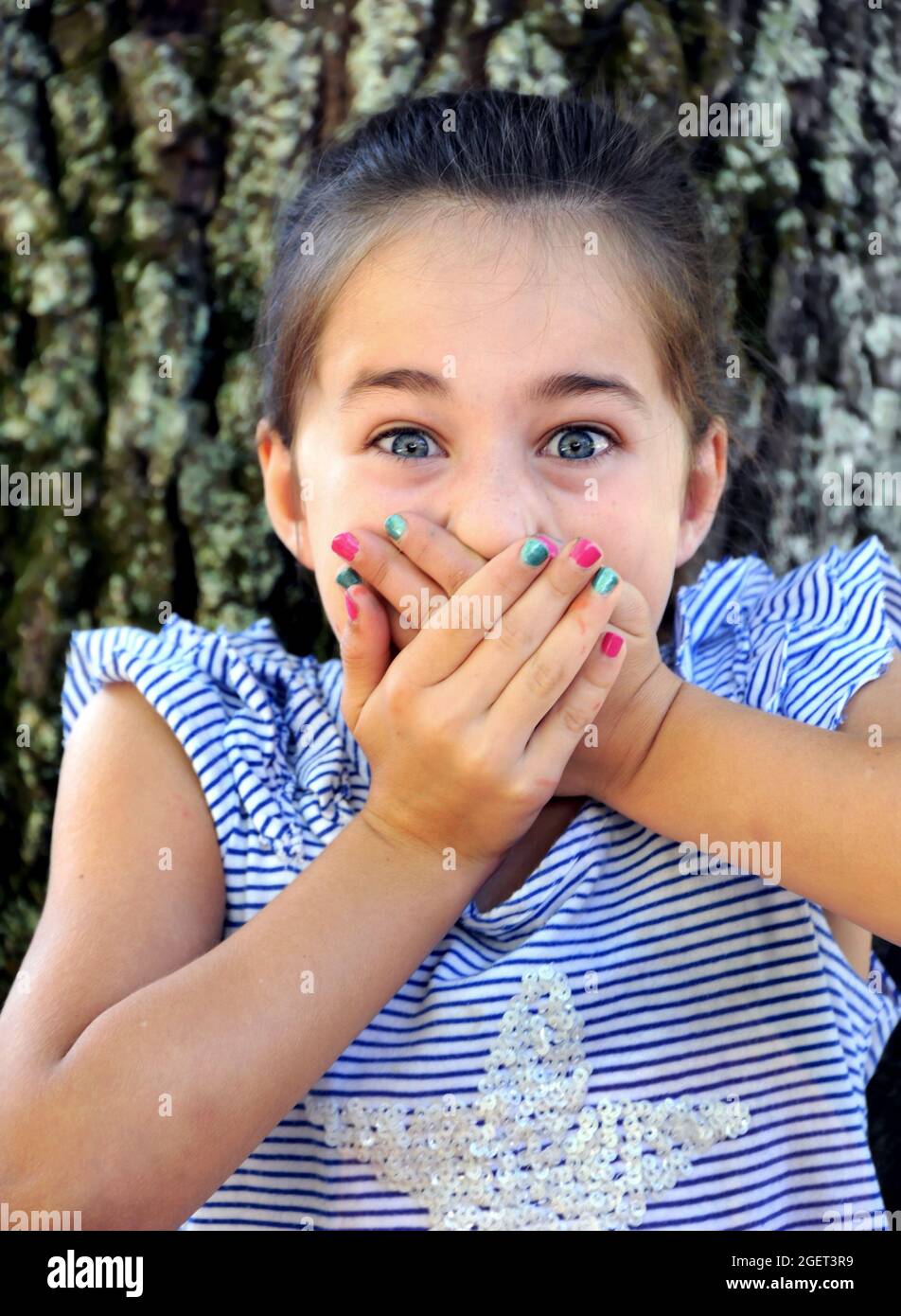 Das kleine Mädchen hält sich in ihrem Überraschungsschrei fest, während sie ihre Hände über ihren Mund hält. Sie sitzt draußen an einem Baum, und das Foto ist aus der Nähe. Stockfoto