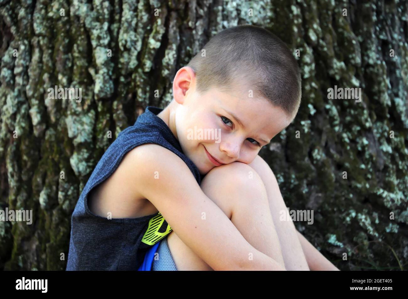 Der kleine Junge legt seinen Kopf gegen seine Knie, während er seitlich auf die Kamera schaut. Er sitzt draußen neben einem großen Baum. Stockfoto