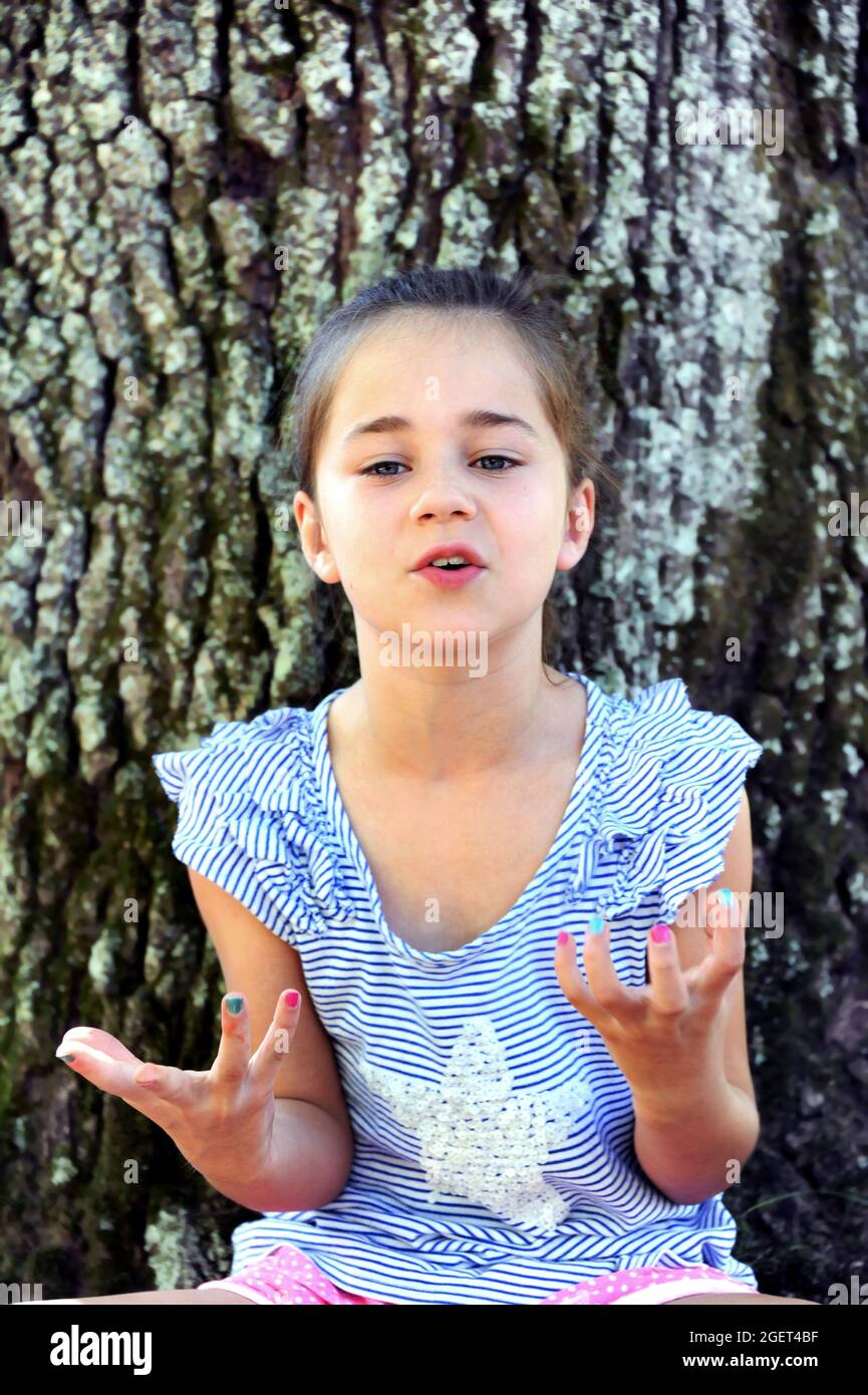 Das kleine Mädchen, das draußen an einem Baum sitzt, ist sehr frustriert und fragt sich, warum sie etwas tun muss. Ihre Hände greifen und sie nutzt Theatralik Stockfoto