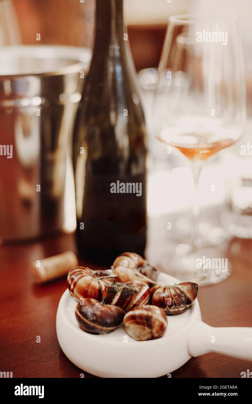 Vertikale Aufnahme von Escargots auf weißer Keramikpfanne serviert neben Weißwein, köstlichen gekochten Schnecken, genießen traditionelle französische Küche im Restaurant Stockfoto