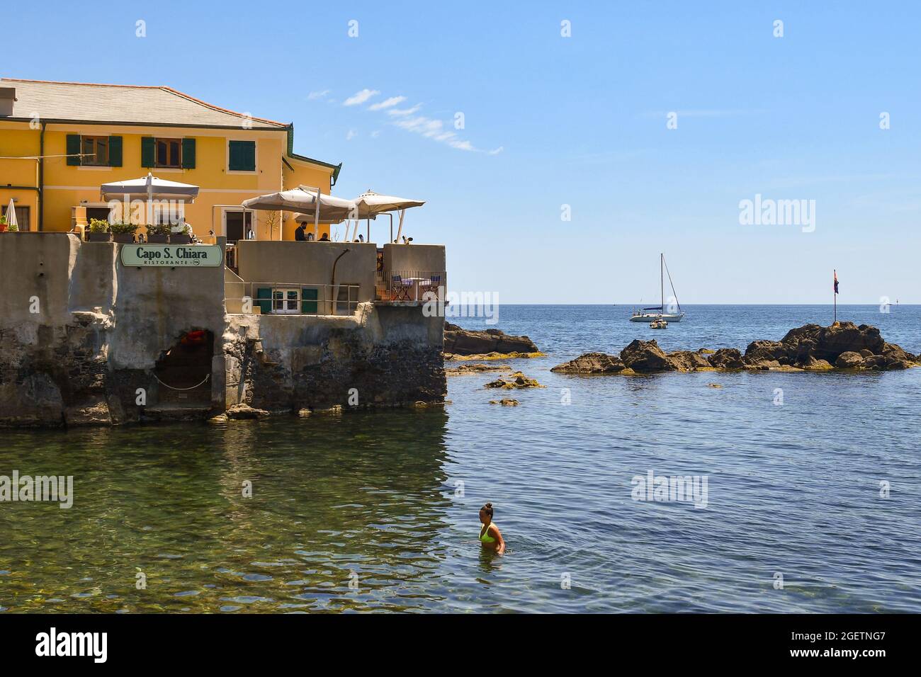 Eine Frau erfrischt sich an der Küste des kleinen Fischerdorfes, während Touristen auf der Terrasse eines Restaurants, Boccadasse, Genua, Italien, speisen Stockfoto