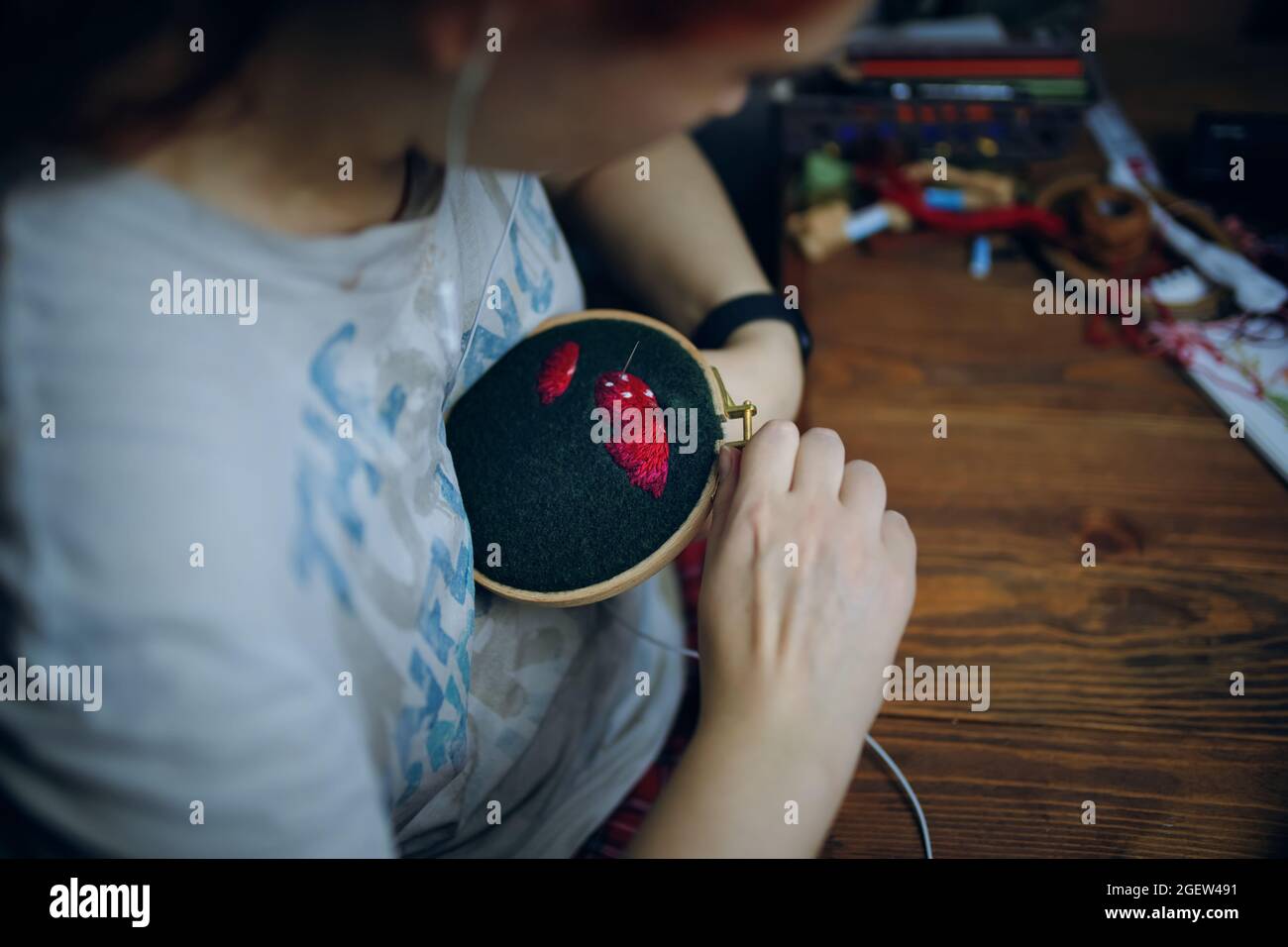 Mädchen stickt Pilzmütze auf grünem Tuch. Handarbeit, Handwerk, Hobby. Frau, die zu Hause auf einem Holzrahmen Zahnseide mit Fäden näht. Stockfoto