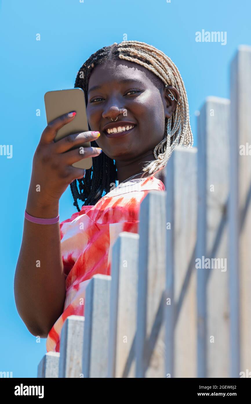 Frau telefoniert und schaut mit einem Holzzaun vor ihr und einem blauen Himmel dahinter auf die Kamera Stockfoto