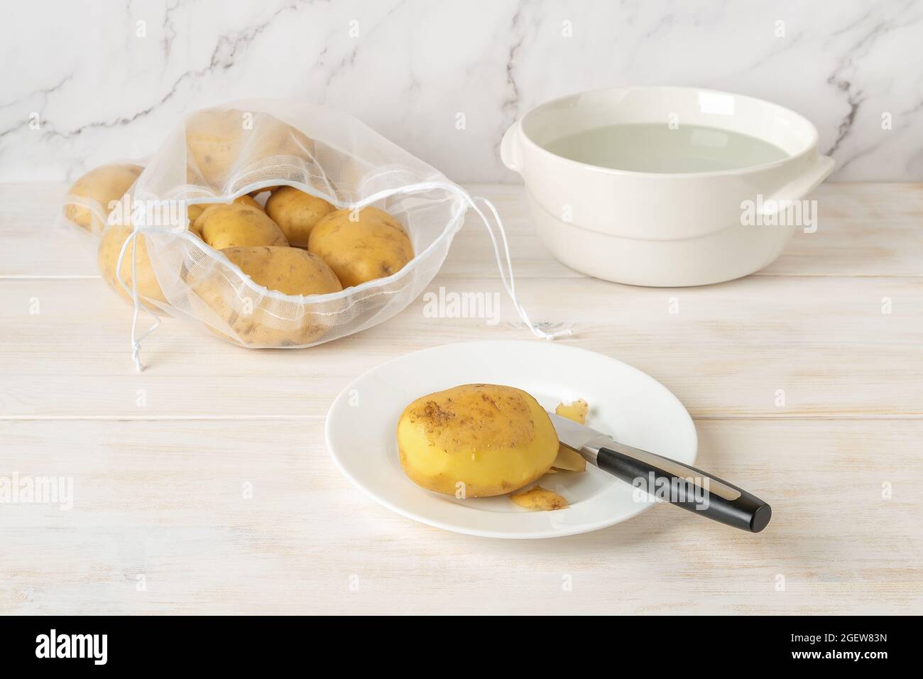 Halb geschälte rohe Kartoffelknolle und Messer auf einem Teller und wiederverwendbarer Nylon-Beutel mit ungeschälten Bio-Kartoffeln über weißem Holz Küchentisch. Stockfoto