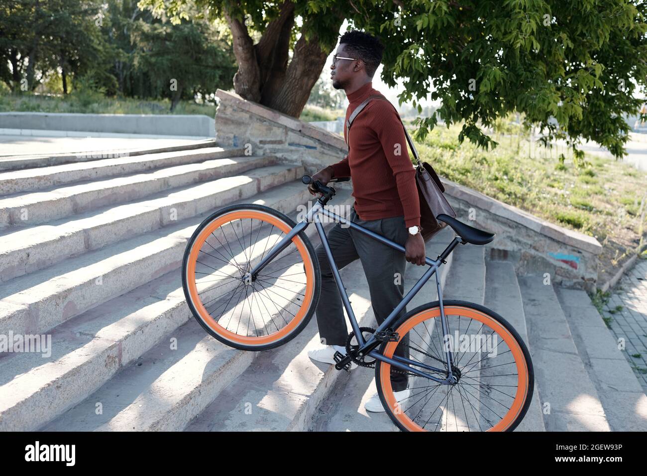Seitenansicht eines jungen afrikanischen Geschäftsmannes, der an einem sonnigen Tag das Fahrrad schiebt, während er nach oben fährt Stockfoto