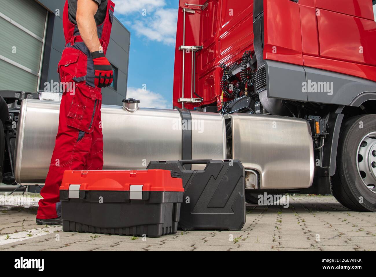 Kaukasischer Semi-Truck-Mechaniker trägt rote Uniform vor dem roten Fahrzeug erforderliche Wartung. Seine professionellen Werkzeugkästen liegen auf einem Boden. Stockfoto