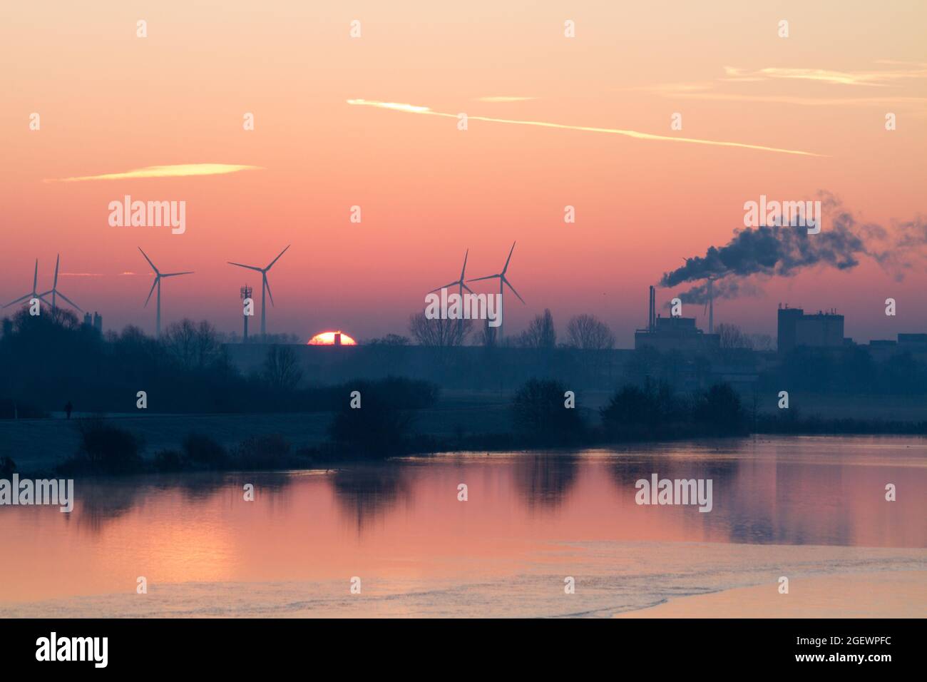 Sonnenaufgang hinter Windkraftanlagen und ein Rauchschornstein mit Rauch, der die Luft verschmutzt, symbolisiert den Klimawandel und den Bedarf an sauberer Energie Stockfoto