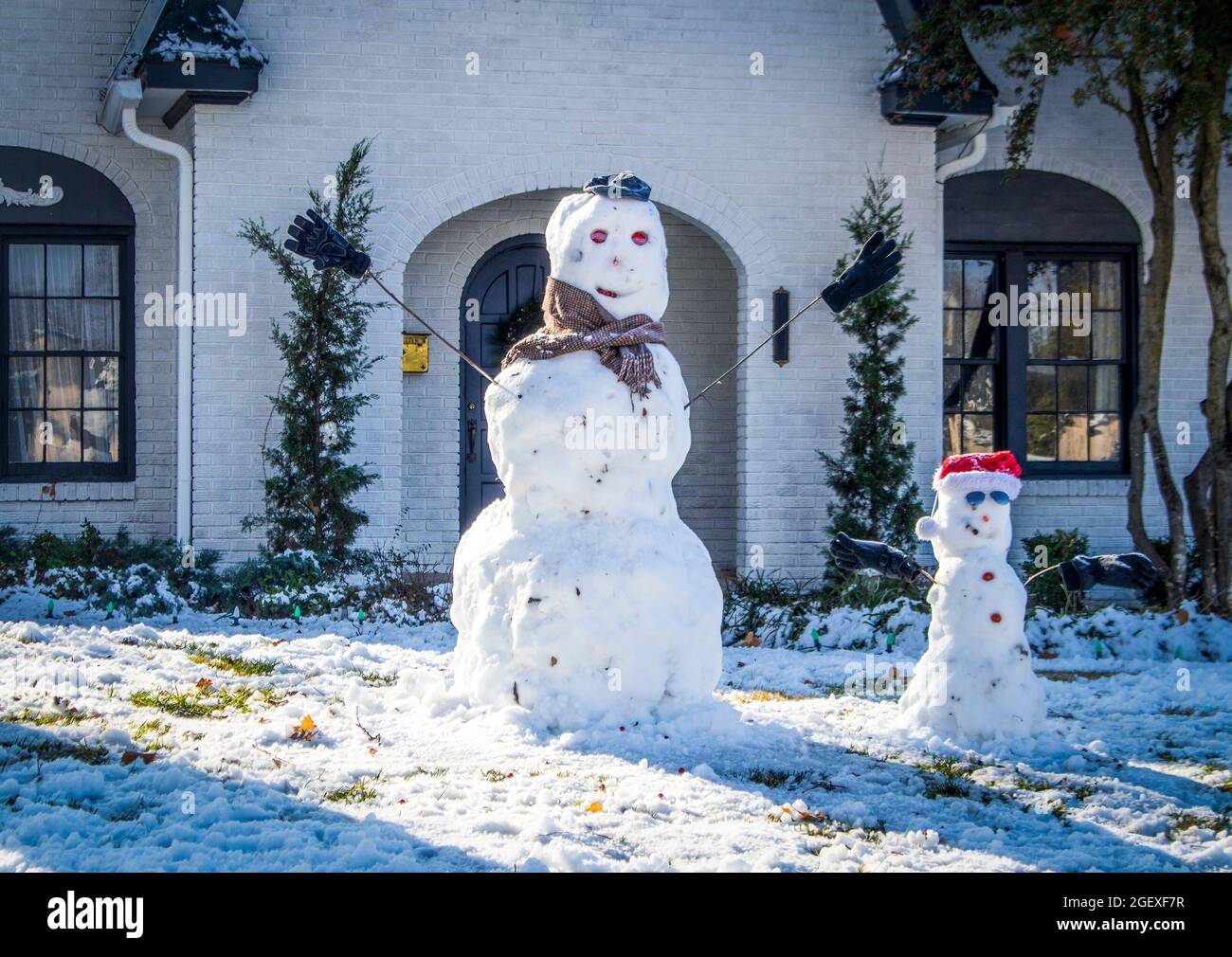 Zwei niedliche Schneemänner vor einem weiß gestrichenen Backsteinhaus - ein großes mit Schal und ein kleines mit weihnachtsmütze Stockfoto