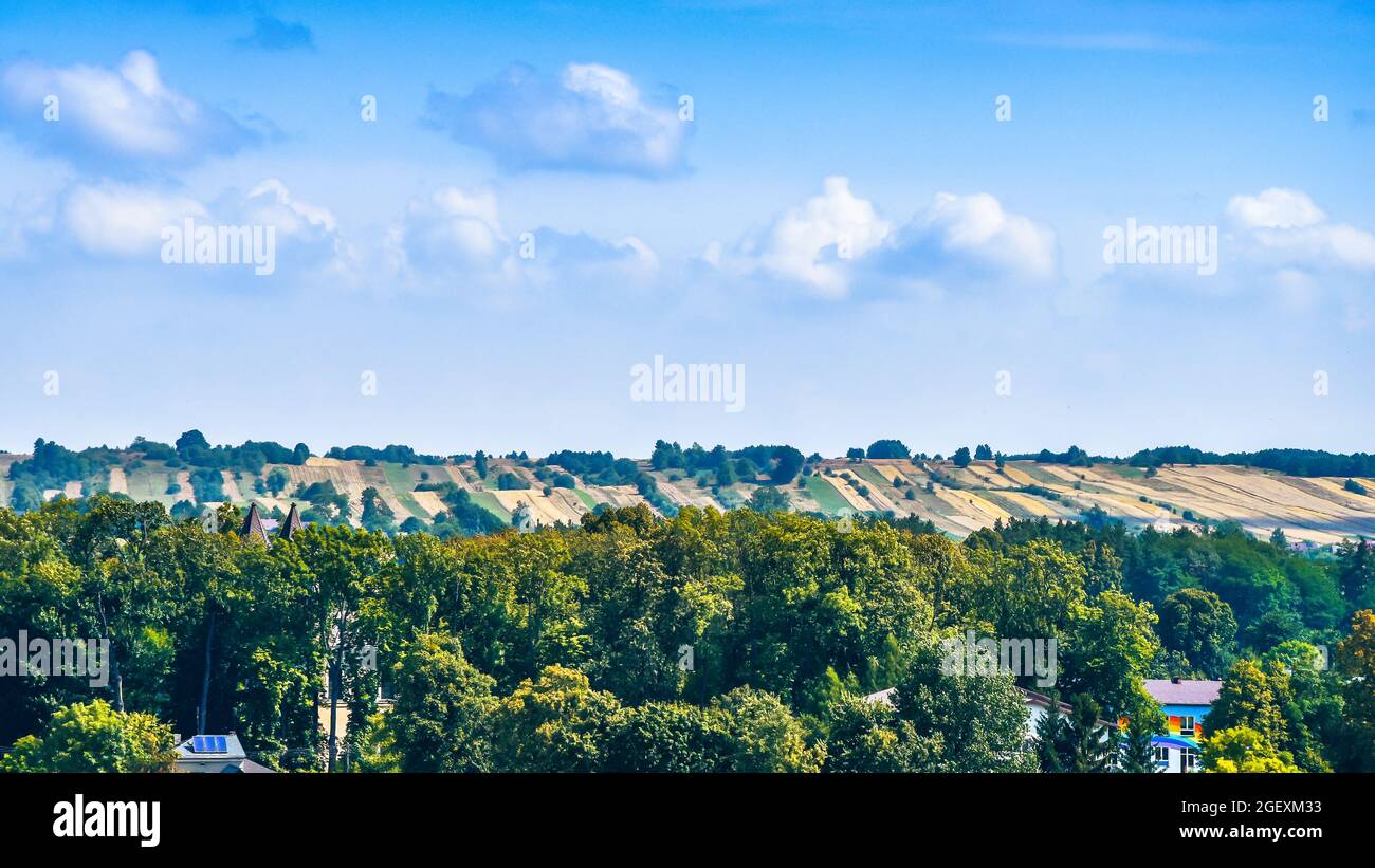 Hügel bedeckt mit landwirtschaftlichen Feldern an sonnigen Sommertagen. Bunte Gebäude umgeben von Bäumen im Vordergrund. Roztocze, Polen. Stockfoto