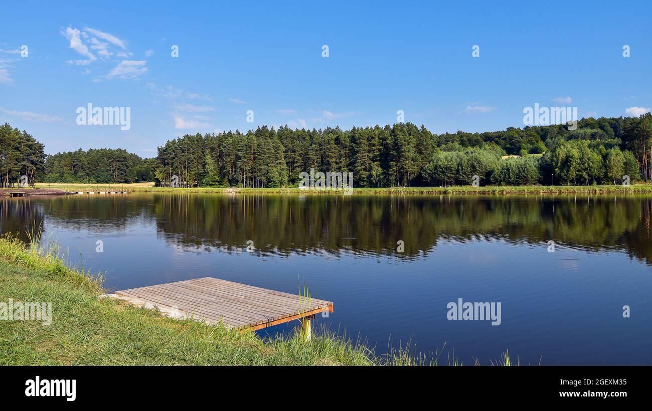 Kleiner Steg, der neben dem See steht. Ruhige und ruhige Oberfläche des Wassers. Hügel mit Wald im Hintergrund bedeckt. Jacnia, Roztocze, Polen. Stockfoto