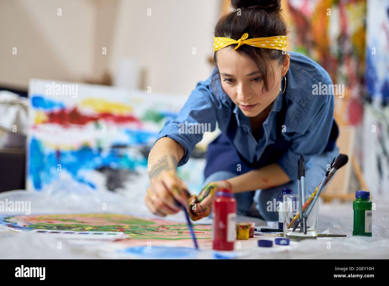Fokussierte junge Frau, Malerin in Schürze Wahl Farbe, halten Pinsel während der Arbeit an der Malerei, sitzen auf dem Boden zu Hause Werkstatt Stockfoto