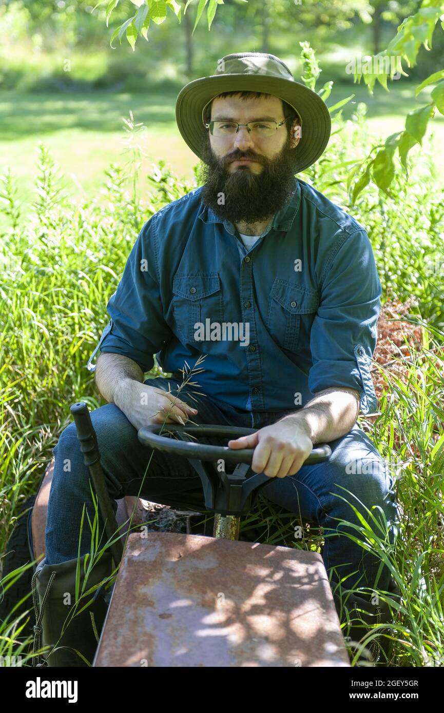 Ein hübscher kaukasischer Rancher, der auf einem alten Grasschneider auf dem Feld sitzt Stockfoto