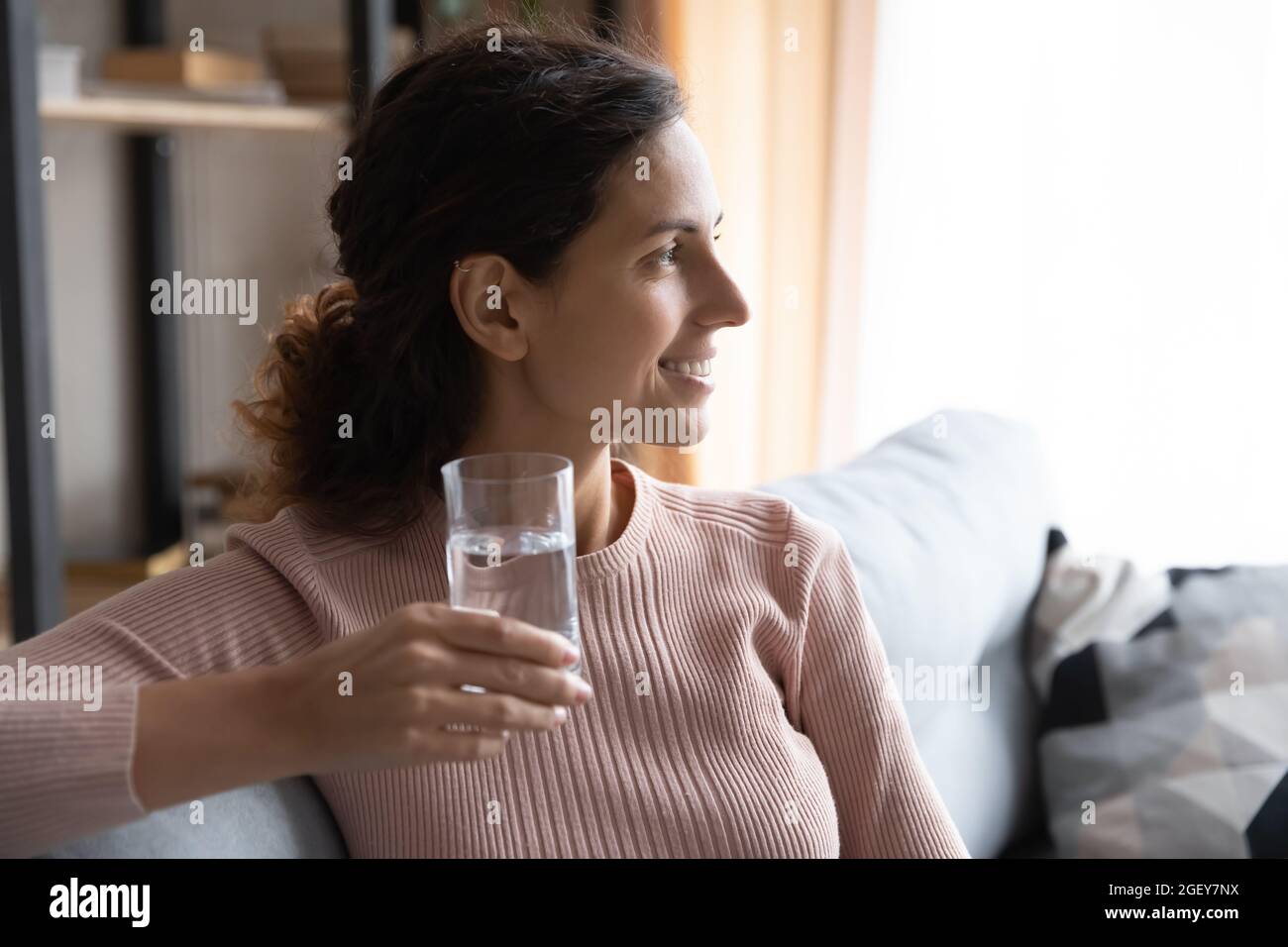 Frau sitzt drinnen und hält ein Glas Wasser in die Ferne Stockfoto