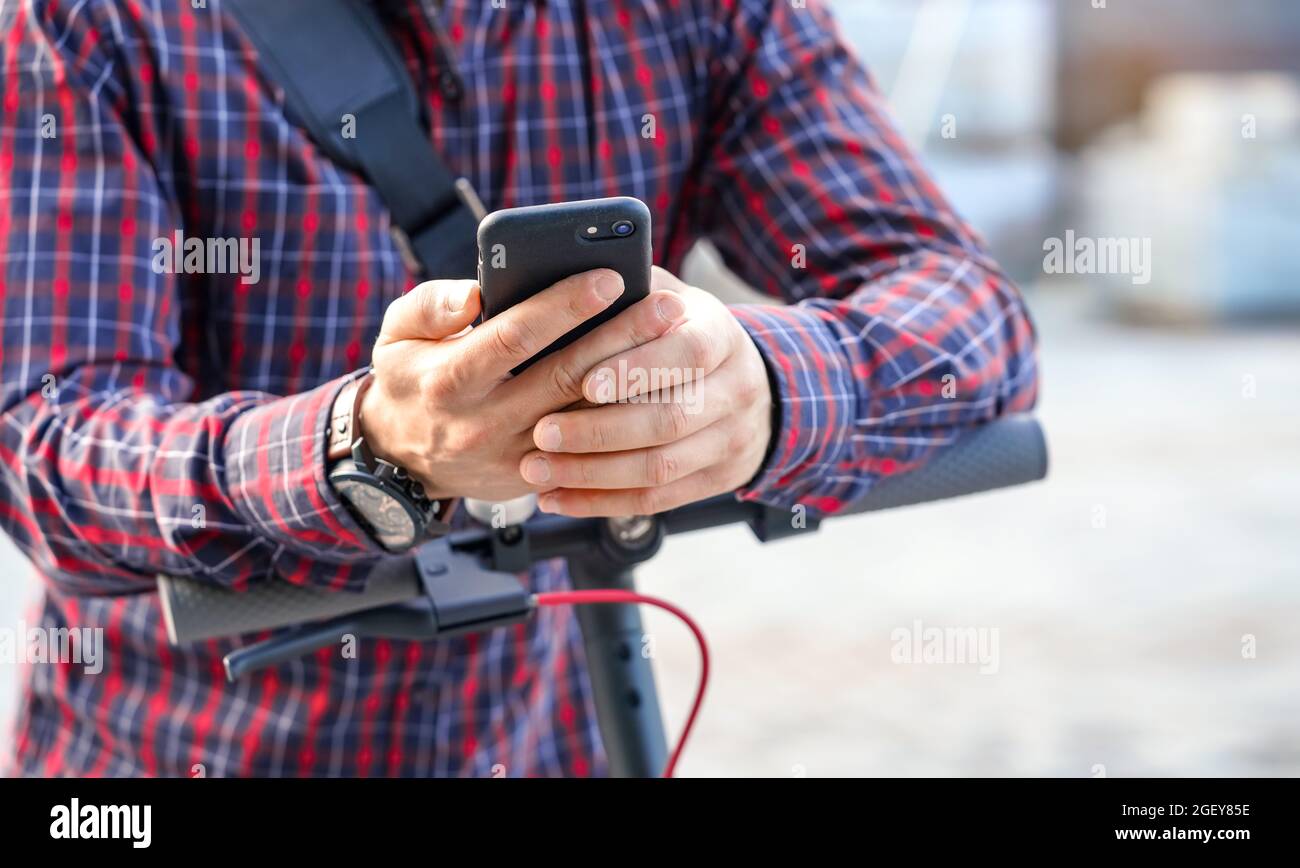 Junger Mann trägt Hemd auf Elektroroller Lenker lehnt, hält Handy in den Händen, Nahaufnahme Detail. Stockfoto