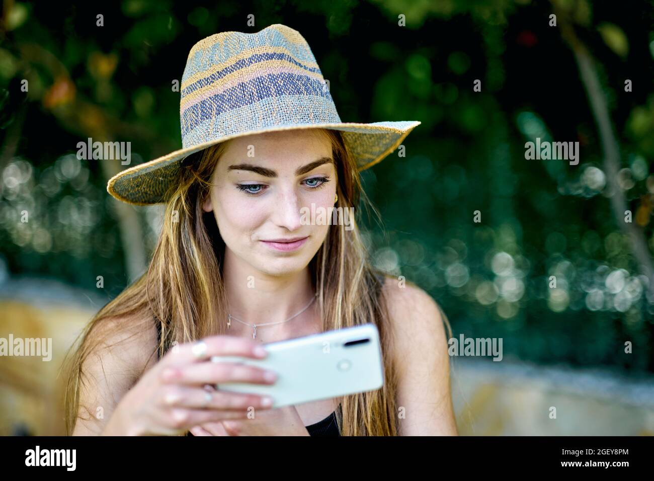 Natürliches Schönheitsporträt einer jungen Frau in den Zwanzigern mit langen Haaren und blauen Augen im Freien in einem Garten, der einen Hut trägt und mit ihrem Handy ein Selfie schießt Stockfoto