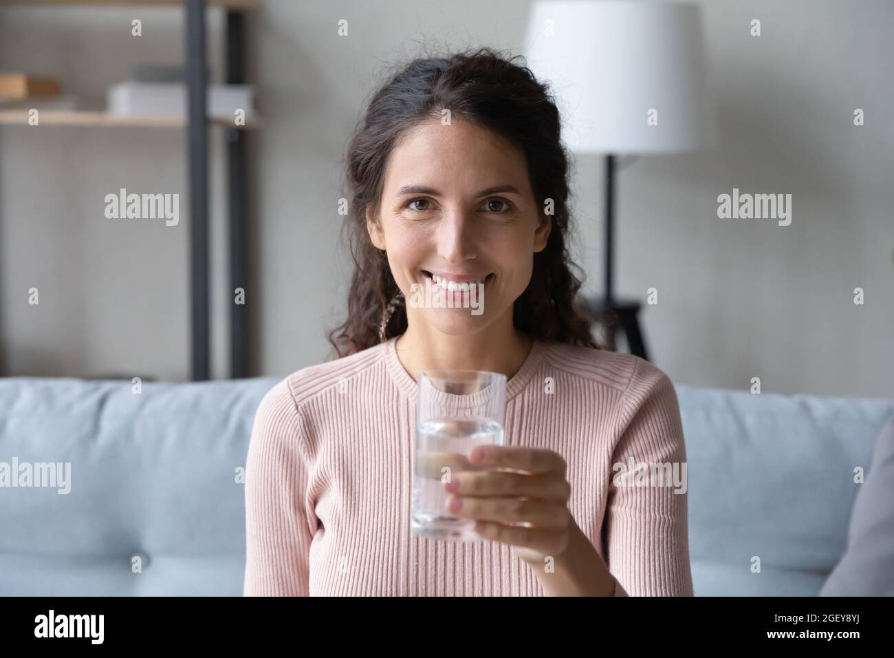 Die junge lateinische Frau sitzt auf dem Sofa und hält ein Glas Wasser Stockfoto